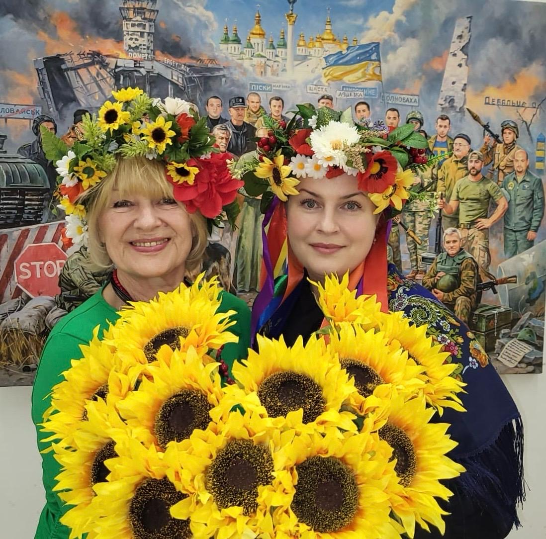 Two women in floral garland crowns holding a large bunch of sunflowers.