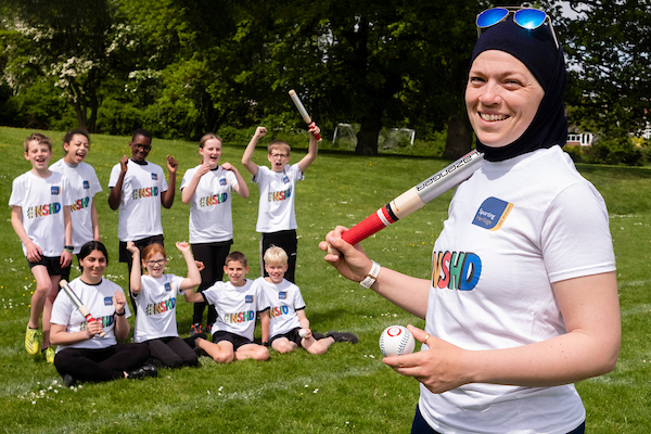 Woman wearing a hijab and carrying a rounders bat and ball, standing in front of a group of schoolchildren on a sports field.