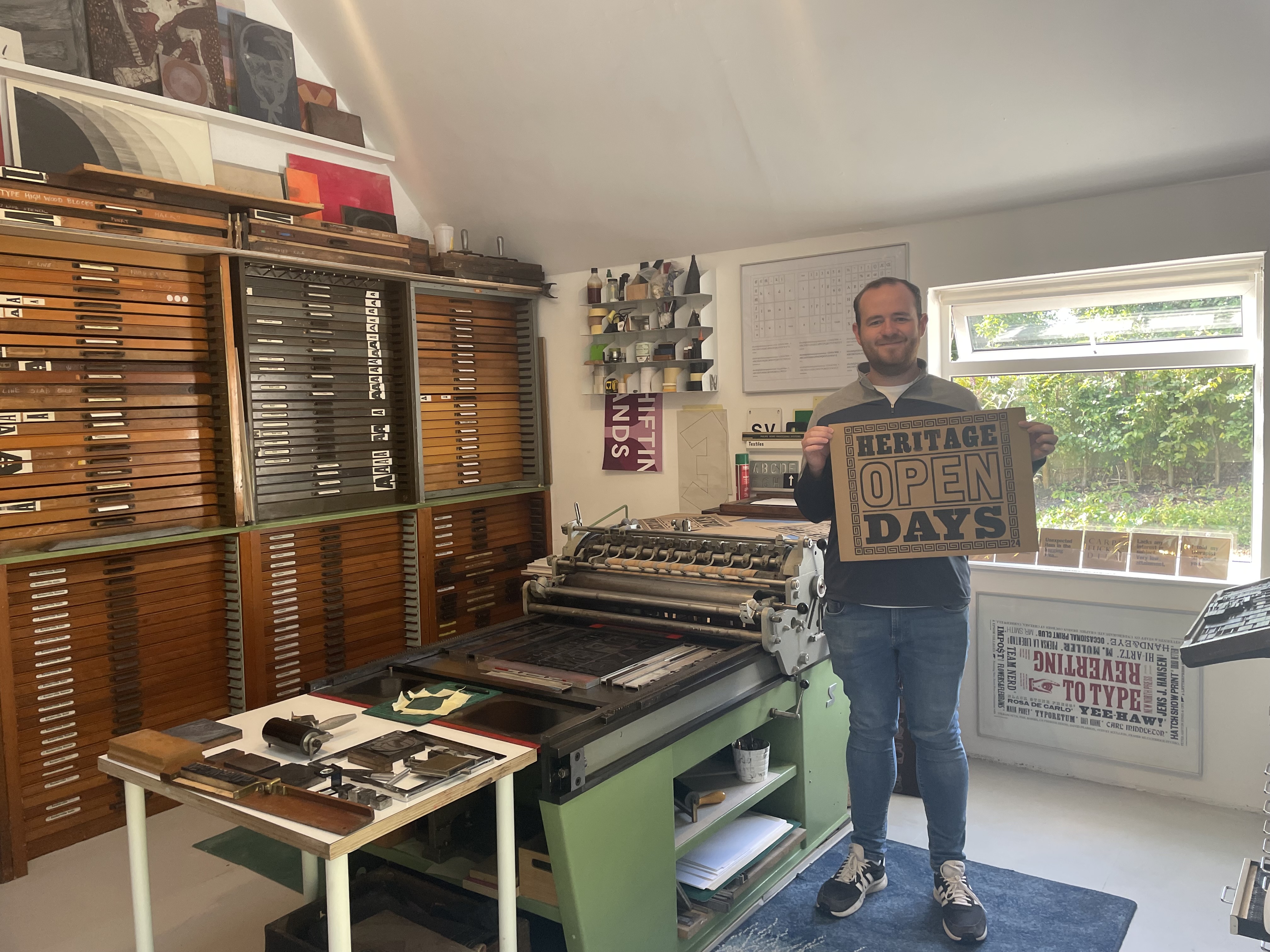 Man standing next to a letter press machine in front of rows of narrow drawers, holding up a printed cardboard sign.