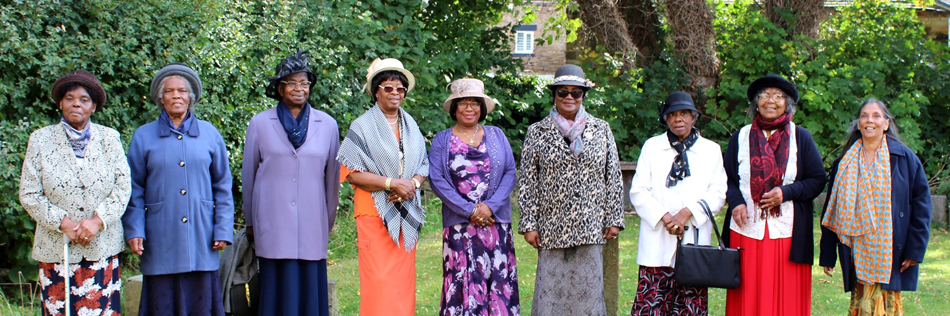 9 black women in formal clothes and hats stood in a line in a garden.