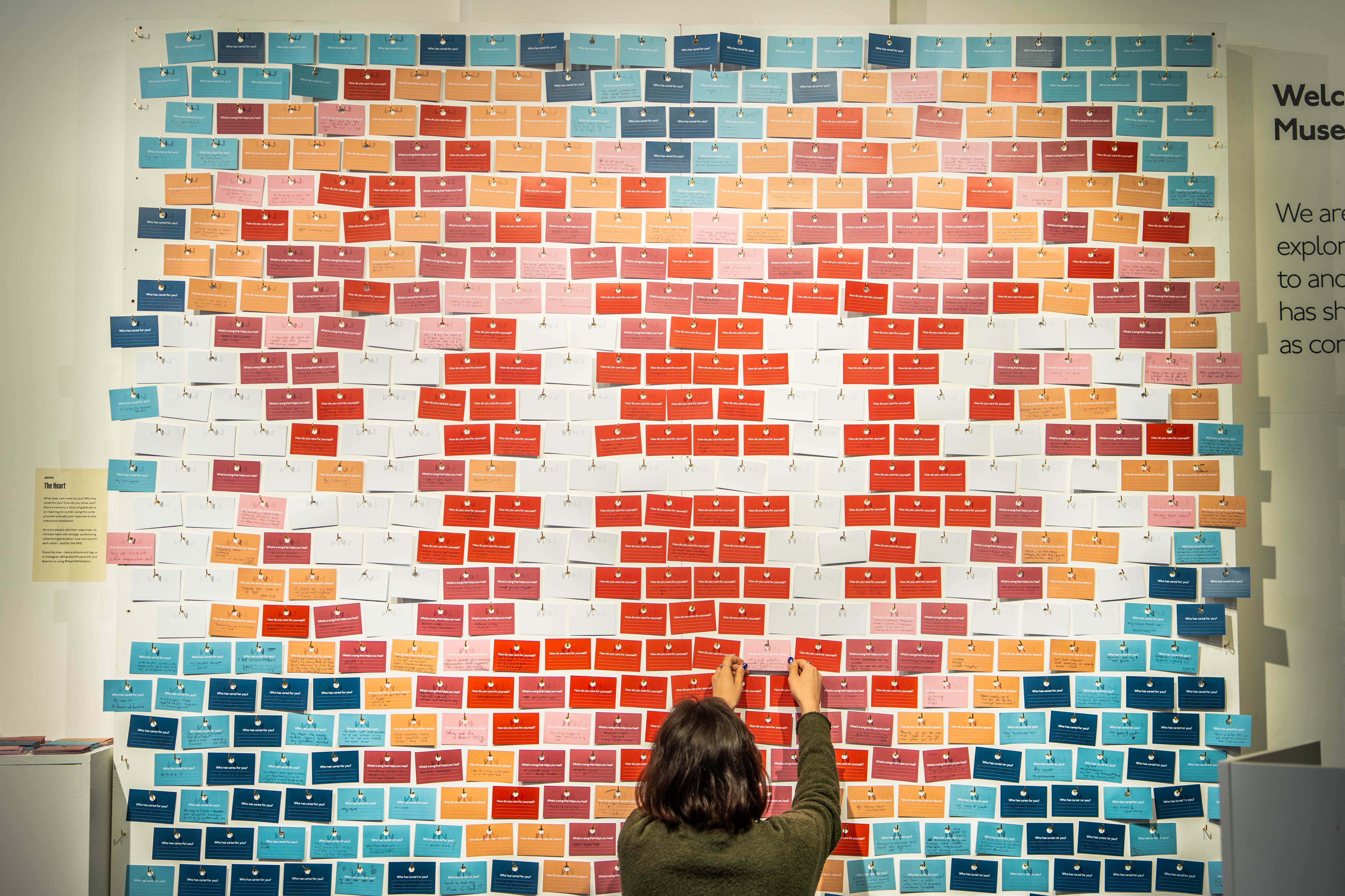 Wall of small rectangular coloured cards pinned into a display of a heart with the letters NHS across it.