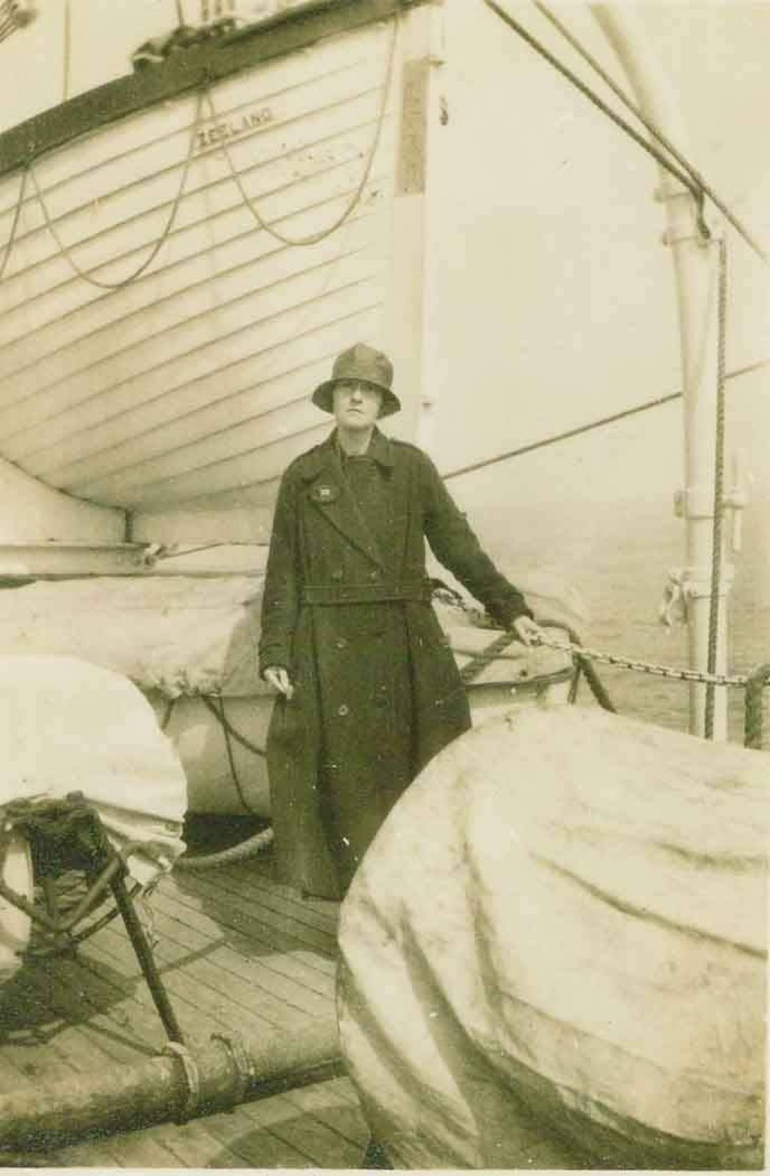 A black and white photograph of a women on a boat. She wears a round hat that covers most her face, a long coat and is stood amongst stored lifeboats.