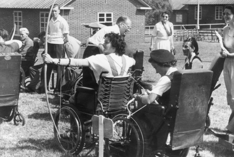 Old photograph of two women archers in wheelchairs on a grass field with a line of people watching behind them.