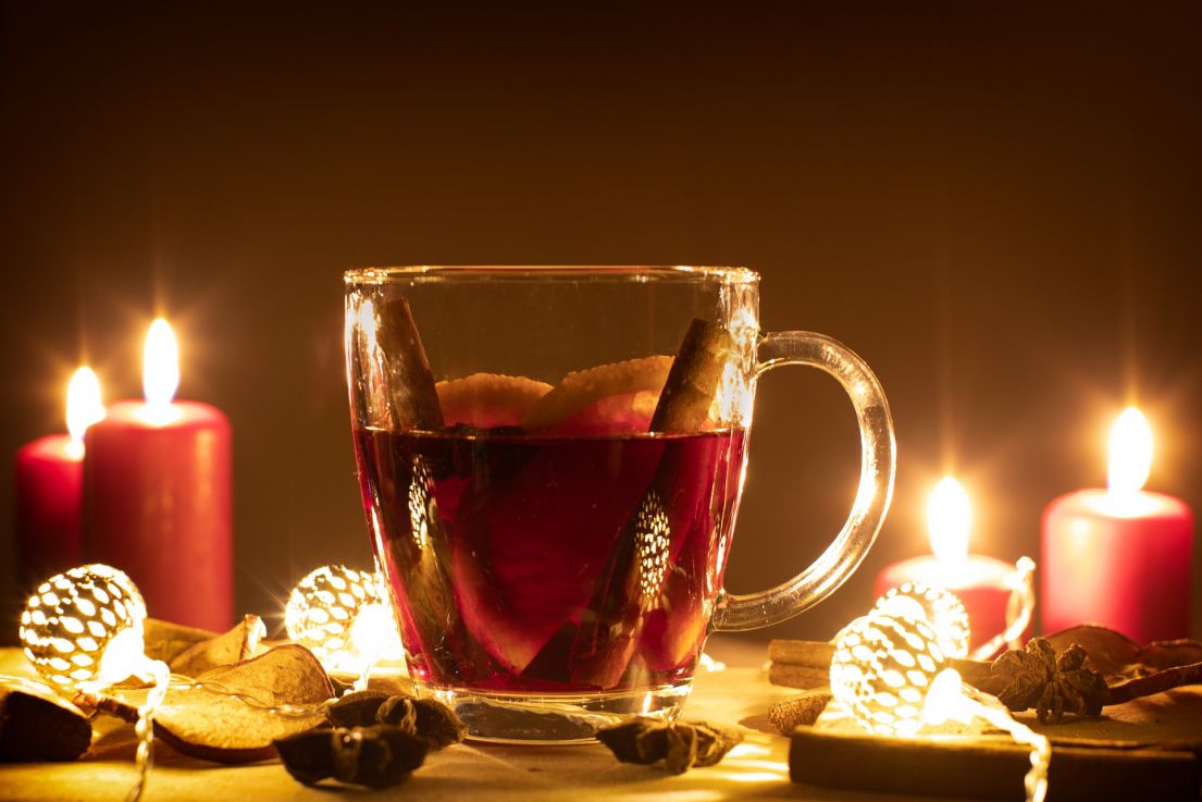 A glass mug of a red drink, with orange slices and cinnamon sticks. Around the mug are fairly lights and candles.