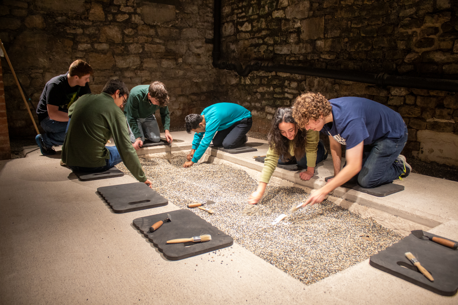 A group of six people kneeling around an indoor archaeological dig pit.