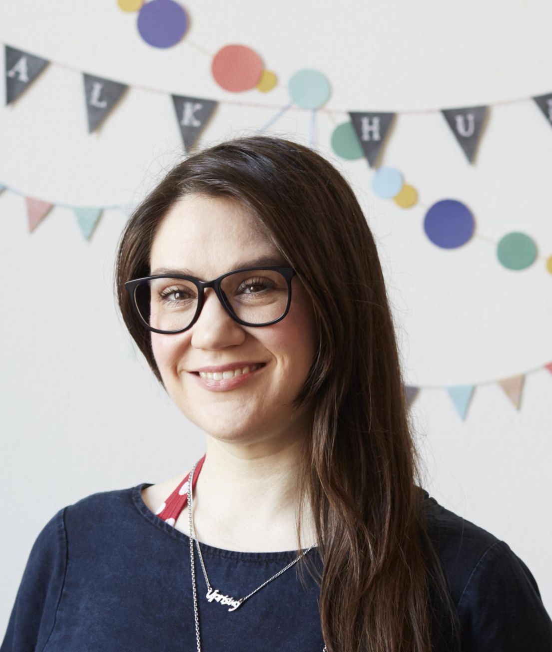 A woman wearing glasses, a blue top, long brown hair,  smiling. Behind her is bunting.