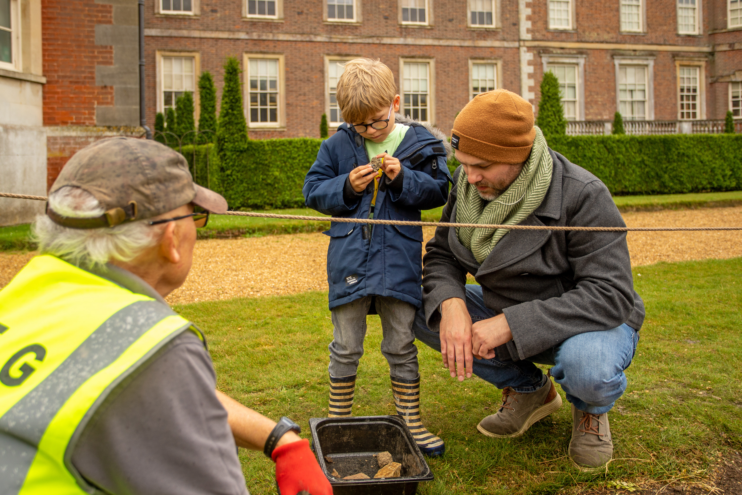 Older man in high vis showing a younger man and child a small object. Knelt on ground by a pit in front of a grand house.