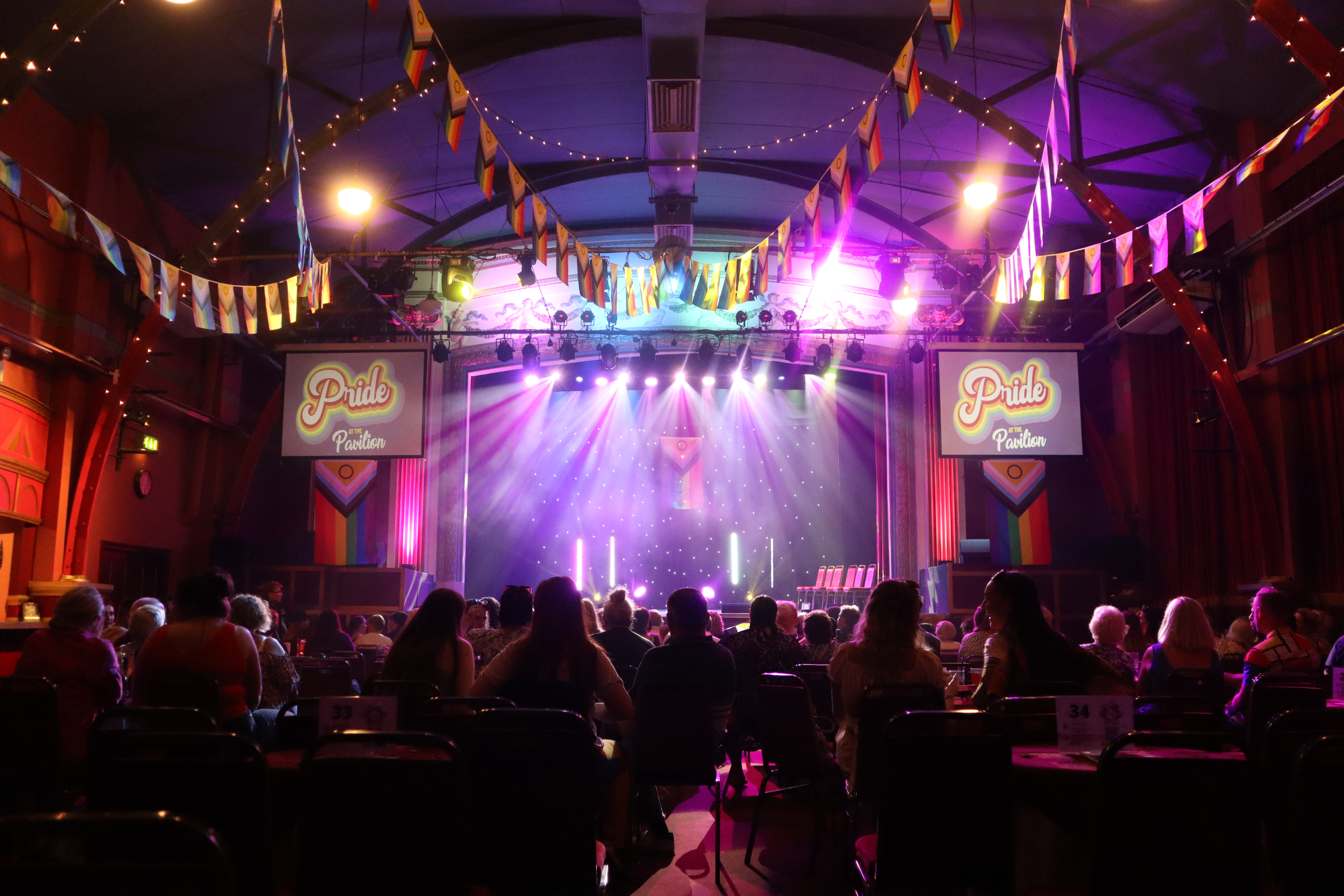 A crowd looking towards a theatre stage with multi-colour lighting, pride flags, and bunting decorating on the ceiling.