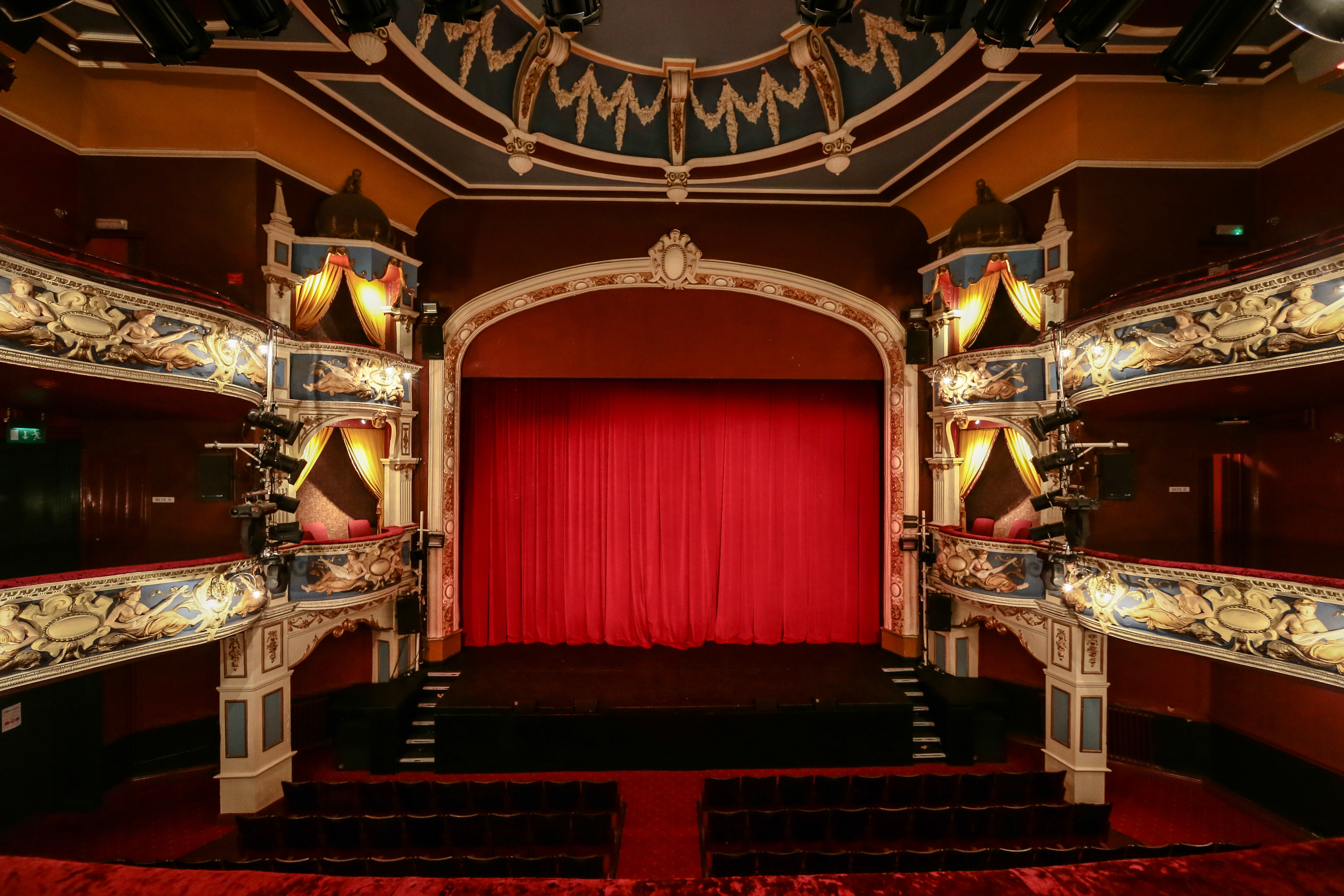 Ornate theatre auditorium with 2 tiers of gilded galleries and a red velvet curtain across the stage.