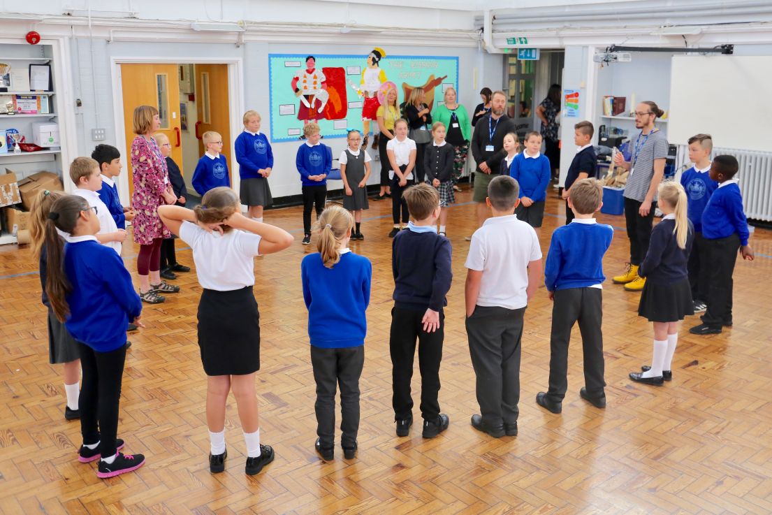 Primary school children with blue uniform jumpers, stood in a circle in a wooden floored room.