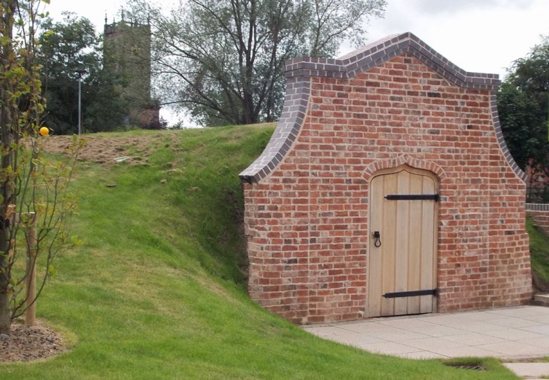 Brick frontage with wooden door backing into a small grass mound with a church tower amongst trees in the background.