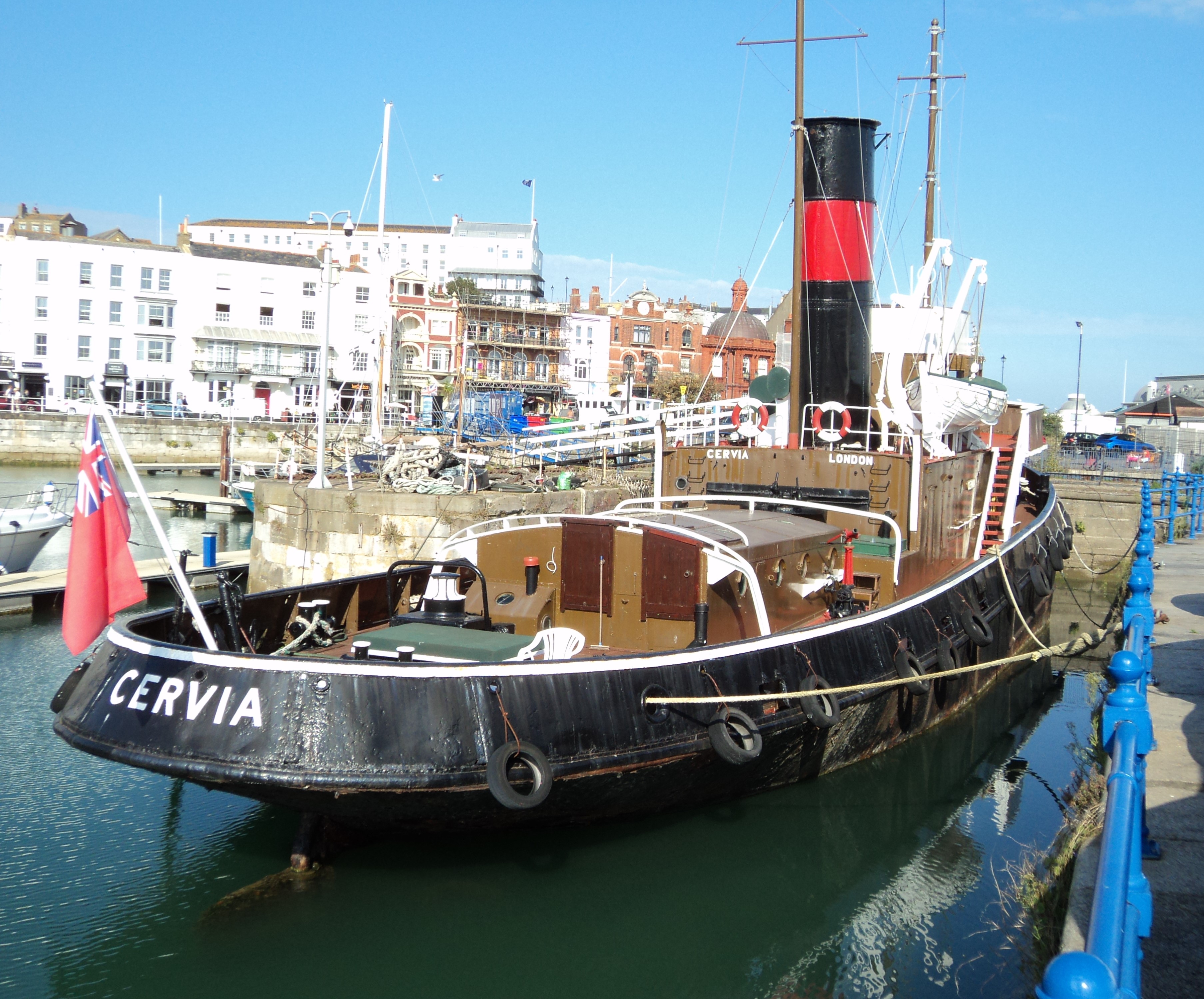 Black hulled boat with chimney docked in a small patch of water with white buildings in the background.