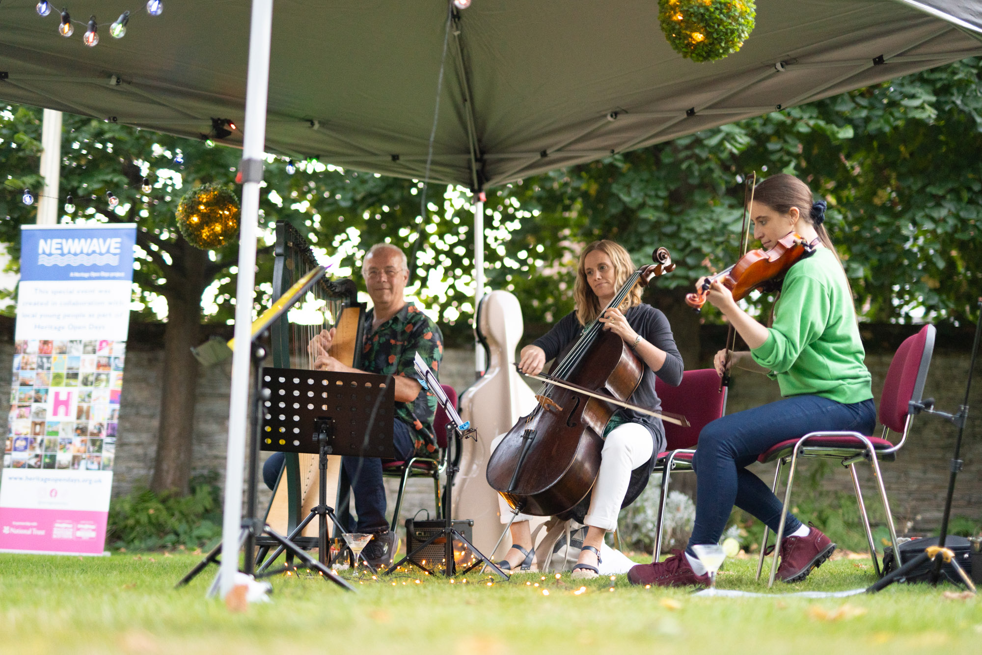 Three people sat on chairs outdoors playing musical instruments - a harp, a cello, and a violin.