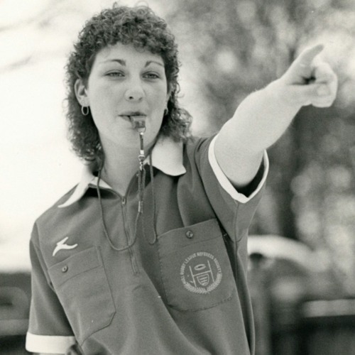 Monochrome photo of a woman in a referee uniform blowing a whistle and pointing.