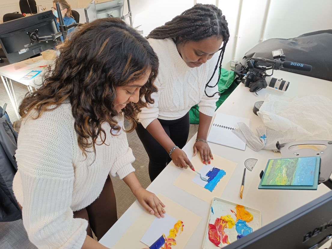 two women with dark hair bent over a table with a small triangular paint spatula, trying to recreate an image in front of them on pieces of paper.