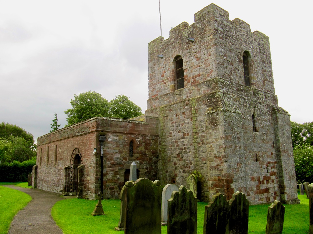 Small brick church with short, squat tower in an area of green grass with trees behind.