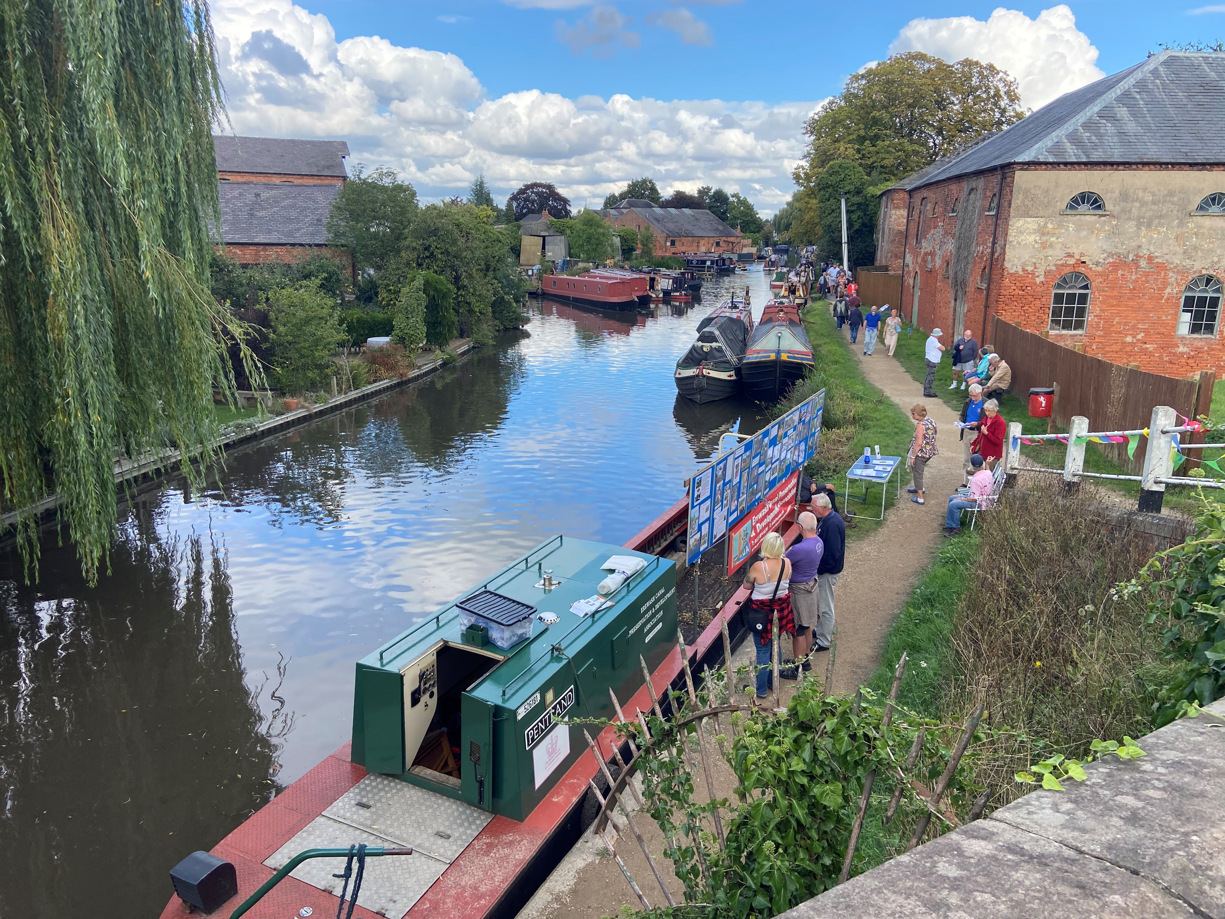 Birds eye view of narrowboats on a canal. Willows on one bank, busy path and a building on the other.