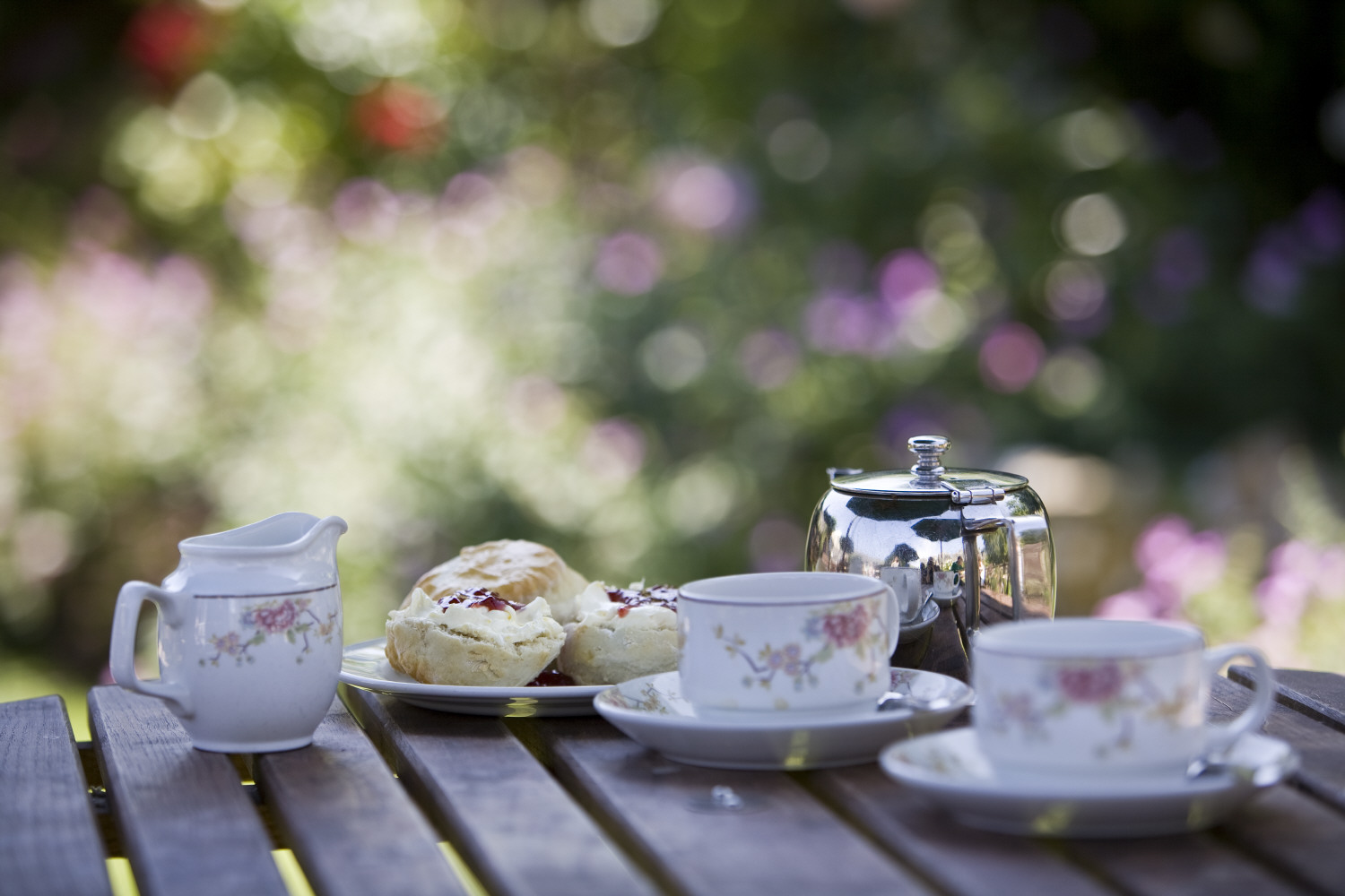 China milk jug, cups and saucers with metal tea pot and plate of scones on a table outside.