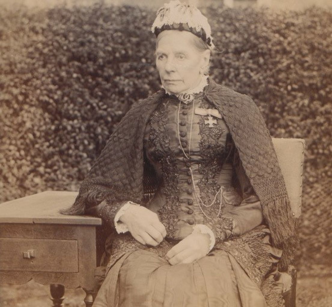 A black and white image of a women in a Victorian dress, her hair is tied up and a Red cross medal is pinned to her chest.