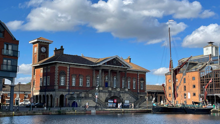 Sunlit harbour with a sailing barge moored in front of an imposing brick building with colonnaded front.